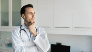 Doctor in white coat with stethoscope around his neck stands in modern medical office, deep in thought and contemplation.